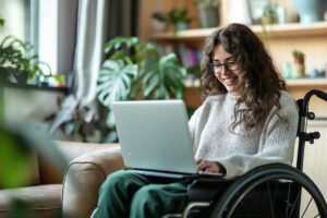 Femme souriante en fauteuil roulant utilisant un ordinateur pour consulter son dossier AAH 2026.
