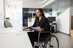 Une femme en fauteuil roulant sourit tout en travaillant sur un ordinateur portable dans un bureau moderne pour illustrer la prime d'activité du travailleur handicapé.